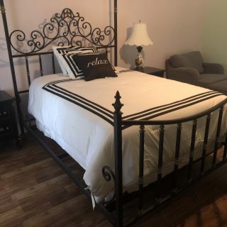 A bedroom featuring an iron bed frame with white bedding and a decorative pillow, accompanied by a lamp and seating area.