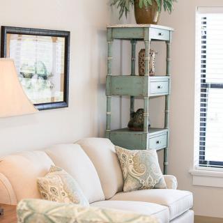 A cozy living room with a white sofa, decorative pillows, and a tall shelving unit with plants near a window.