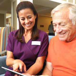 A nurse assists a smiling senior citizen with a tablet.