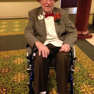 An elderly person dressed in formal attire sitting in a wheelchair indoors.