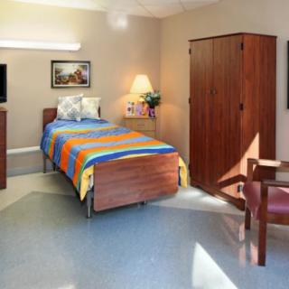 A neatly arranged hospital room with a colorful bedspread, wardrobe, chair, and television.