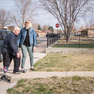 A caregiver assists an elderly person while walking along the sidewalk on a sunny day.