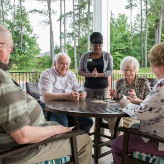 A group of seniors playing cards on a patio with a staff member standing by.