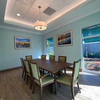 A modern dining or conference room with a large table, surrounded by eight chairs, featuring artwork on the walls and natural light from the windows.