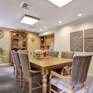 A cozy dining room with a large wooden table and upholstered chairs, softly lit by overhead lights.
