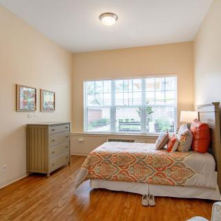 A cozy bedroom with a large window providing natural light, featuring a bed with patterned bedding and a chest of drawers.