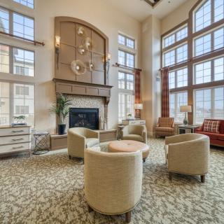 A spacious living room with a high ceiling, featuring large windows, a cozy seating area around a fireplace, and an elegant chandelier.