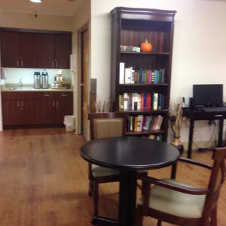 A cozy office space with a round table, bookshelf, and kitchenette in the background.