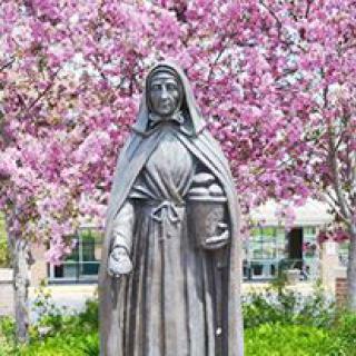 A statue of a cloaked figure stands in front of blooming trees with pink flowers in a garden setting.