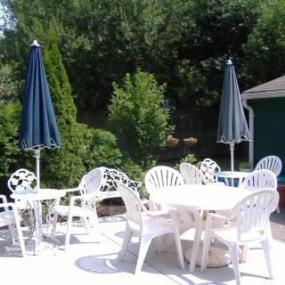 An outdoor patio area with white plastic chairs and tables, shaded by blue umbrellas, surrounded by greenery.