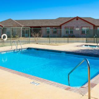 An outdoor swimming pool under a clear blue sky, surrounded by a fence and adjacent to a resort building.