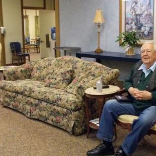 An elderly person sitting in a cozy living room with a floral couch and warm lighting.