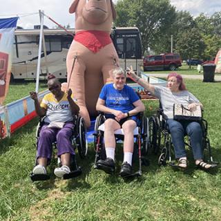 A lively outdoor event featuring people in wheelchairs enjoying their time with a humorous inflatable character behind them.