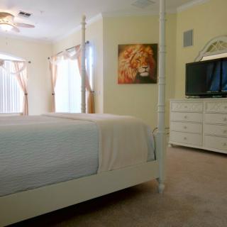 A cozy bedroom with a large bed, dresser, and television, adorned with wall art and natural light streaming through the windows.