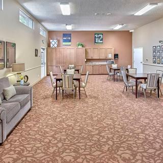 A spacious community room with tables, chairs, and a sofa, featuring a patterned carpet and wall decorations.