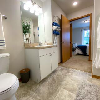 A modern bathroom with a vanity, mirror, and neatly arranged towels leading into a bedroom.