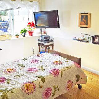A cozy bedroom featuring a floral bedspread, wall-mounted TV, and decorative frames by the window.