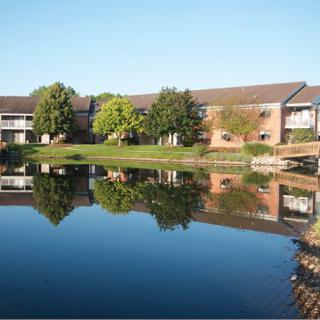 A peaceful residential complex with apartments overlooking a serene lake, reflecting the clear blue sky and surrounding trees.