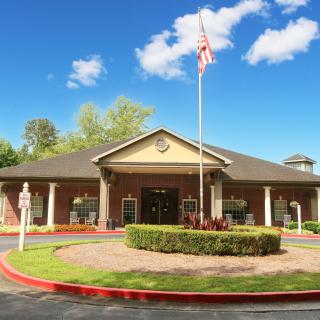 A single-story brick building with a flagpole in front, surrounded by landscaped greenery under a blue sky with clouds.