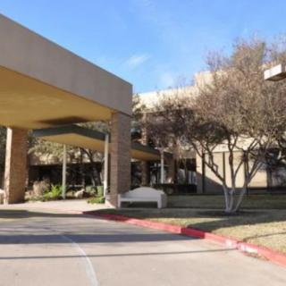 The image shows the entrance of a building with a covered walkway, surrounded by trees and a sidewalk.