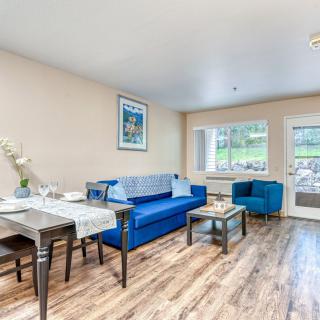 A cozy living room featuring a blue sofa, armchair, dining table, and large window providing natural light.