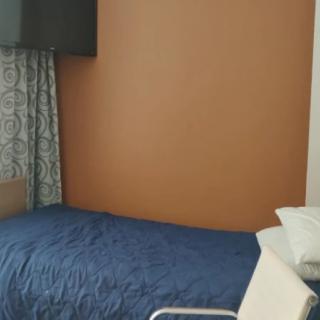 A simple bedroom with a blue bedspread, a pillow, and patterned curtains against an orange wall.