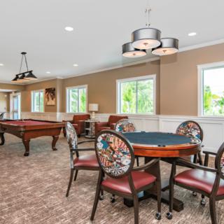A spacious game room featuring a pool table and a poker table with surrounding chairs, well-lit by pendant lights and natural light from large windows.