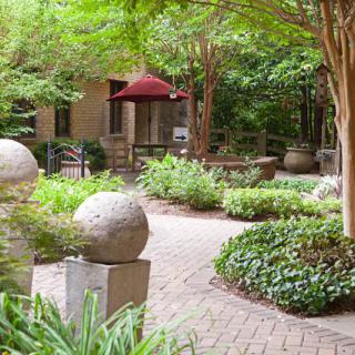 A peaceful garden area with sculptures, greenery, and a winding walkway under trees.