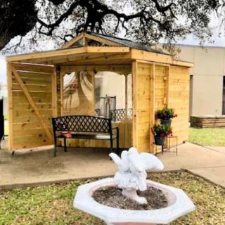 A wooden gazebo with a bench under a tree near a white fountain.