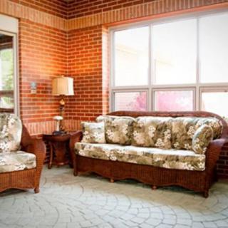 A cozy living room with a floral sofa and armchair against a brick wall, featuring large windows and a lamp.