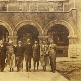 A vintage group photograph of eight individuals standing in front of an ornate building with arches.