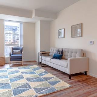 A modern living room with a beige sofa, blue and white rug, and an armchair by the window.