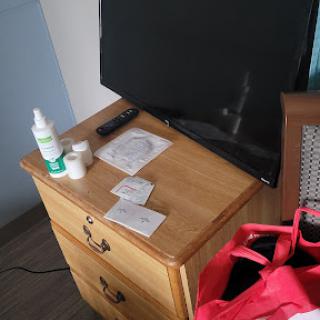 A wooden nightstand in a bedroom with a television, healthcare products, and a pink bag.