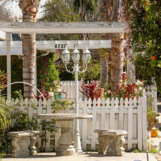 A charming garden featuring a white picket fence, pergola, and outdoor seating surrounded by lush greenery and palm trees.