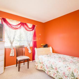 A cozy bedroom with vibrant orange walls, featuring a single bed with floral bedding, a window adorned with curtains, a wooden chair, and a dresser.