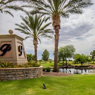 A serene view of a golf course entrance with palm trees, a pond, and lush greenery under a cloudy sky.