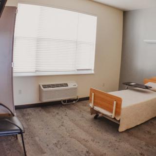 A minimalist hospital room featuring a bed, chair, and air conditioner under a window.