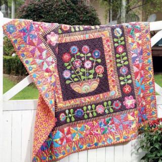 A colorful quilt featuring a floral design is hung outdoors on a white fence.