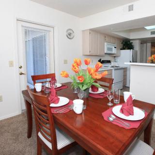 A dining room with a table set for four, featuring vibrant flowers and a view of the kitchen.