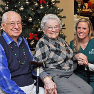 An elderly couple, accompanied by a caregiver, smiles warmly in front of a decorated Christmas tree.