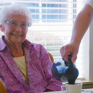 An elderly woman smiling as a person pours coffee into her cup indoors.