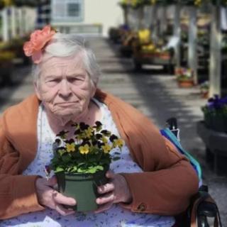An elderly woman in a wheelchair holding a potted flower in a nursery.
