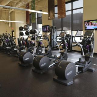 An empty gym with rows of elliptical machines and weightlifting equipment.