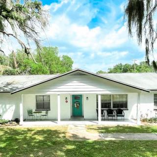 A cozy single-story house with a white exterior and a small front porch, surrounded by lush greenery and under a clear blue sky.