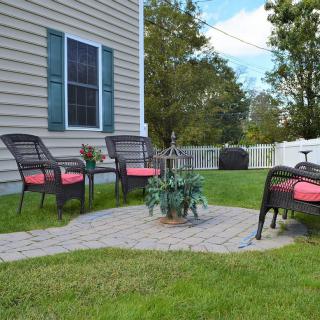 A cozy backyard patio area with wicker chairs and a small table on a paved surface, surrounded by a lawn.