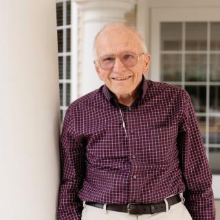 An elderly man smiling indoors, dressed in casual clothing.