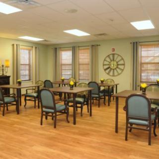 A neatly arranged dining room with multiple tables and chairs, featuring a large wall clock and decorated windows.