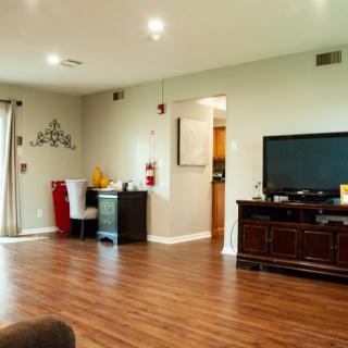 A spacious living room with wooden flooring and minimalist decor, featuring a TV and a desk.