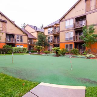A residential apartment complex featuring a well-maintained putting green area surrounded by landscaped greenery.
