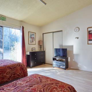 A cozy bedroom with two beds, a dresser, and a television, featuring natural light from a large sliding door.